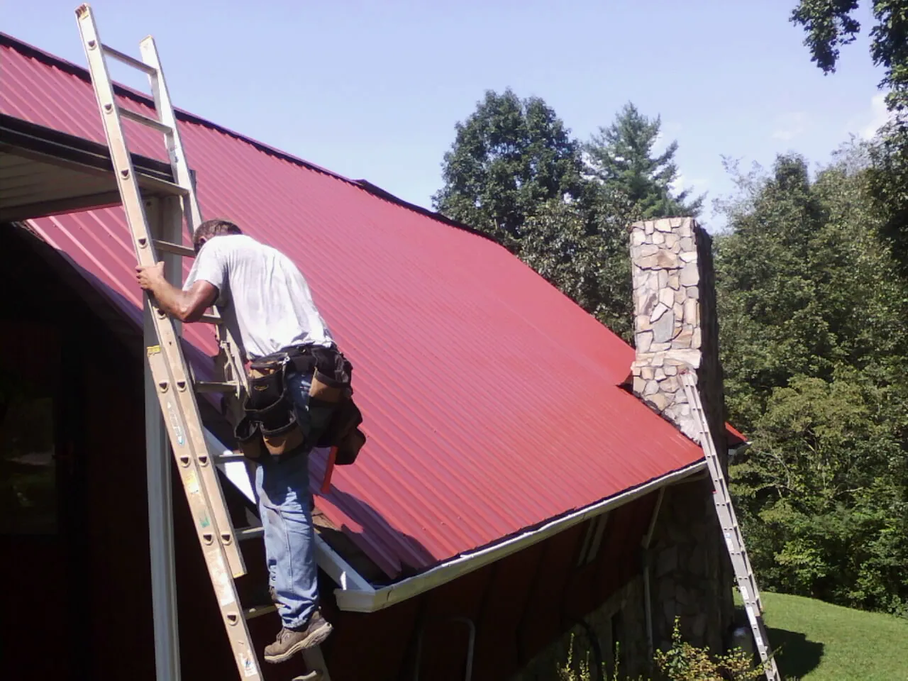 Roofer on a ladder beside a red metal roof and stone chimney — Soffit & Fascia Repair by Roofing Brothers in Glouster